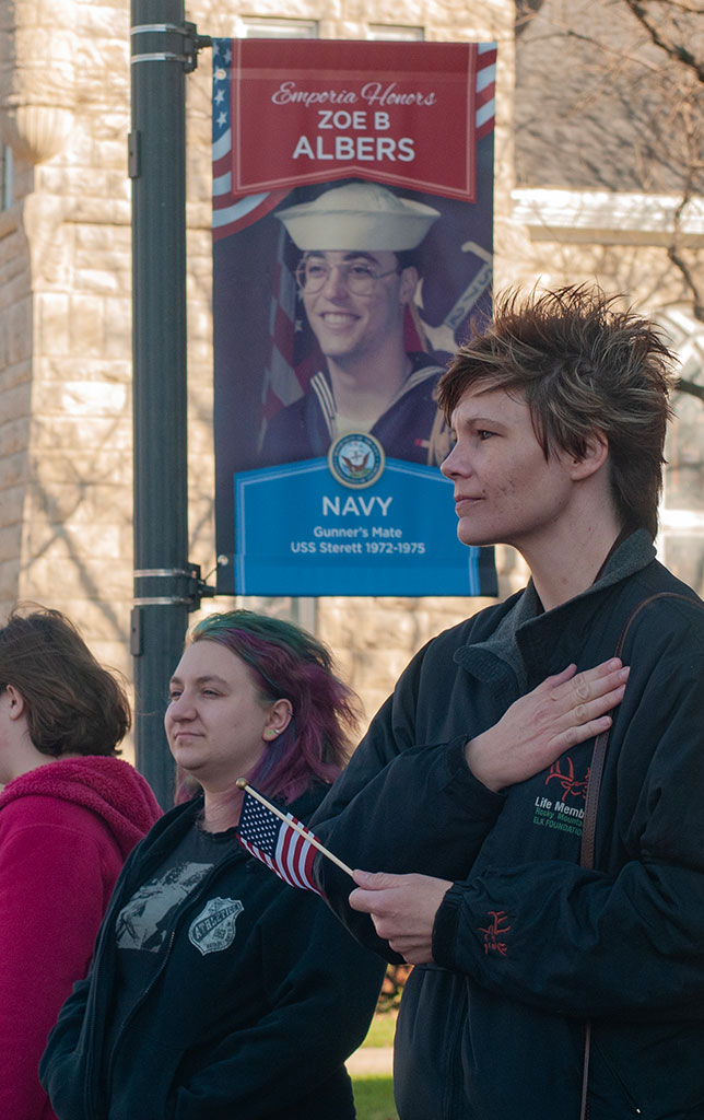 Photo depicting three people standing on Commercial street on Emporia. While The third person in a pink sweater is turned out of frame of the photo, the other two are looking ahead of them. The taller of these two on the right is holding a small American flag in one hand and has their other hand placed over their chest. Behind them is one of the many veteran banners across town, This banner features Zoe B. Albers.